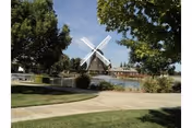 Park-like outdoor scene with a white windmill by a pond, surrounded by trees, lawn, and a paved walkway.