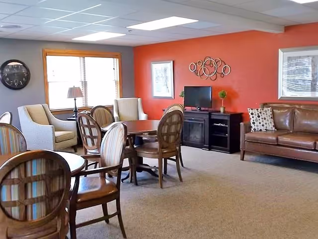 A cozy living room area with a brown leather sofa, several wooden chairs with cushions, two armchairs near a window with blinds, a small table lamp, a TV on a black stand, and decorative wall art on a red accent wall.
