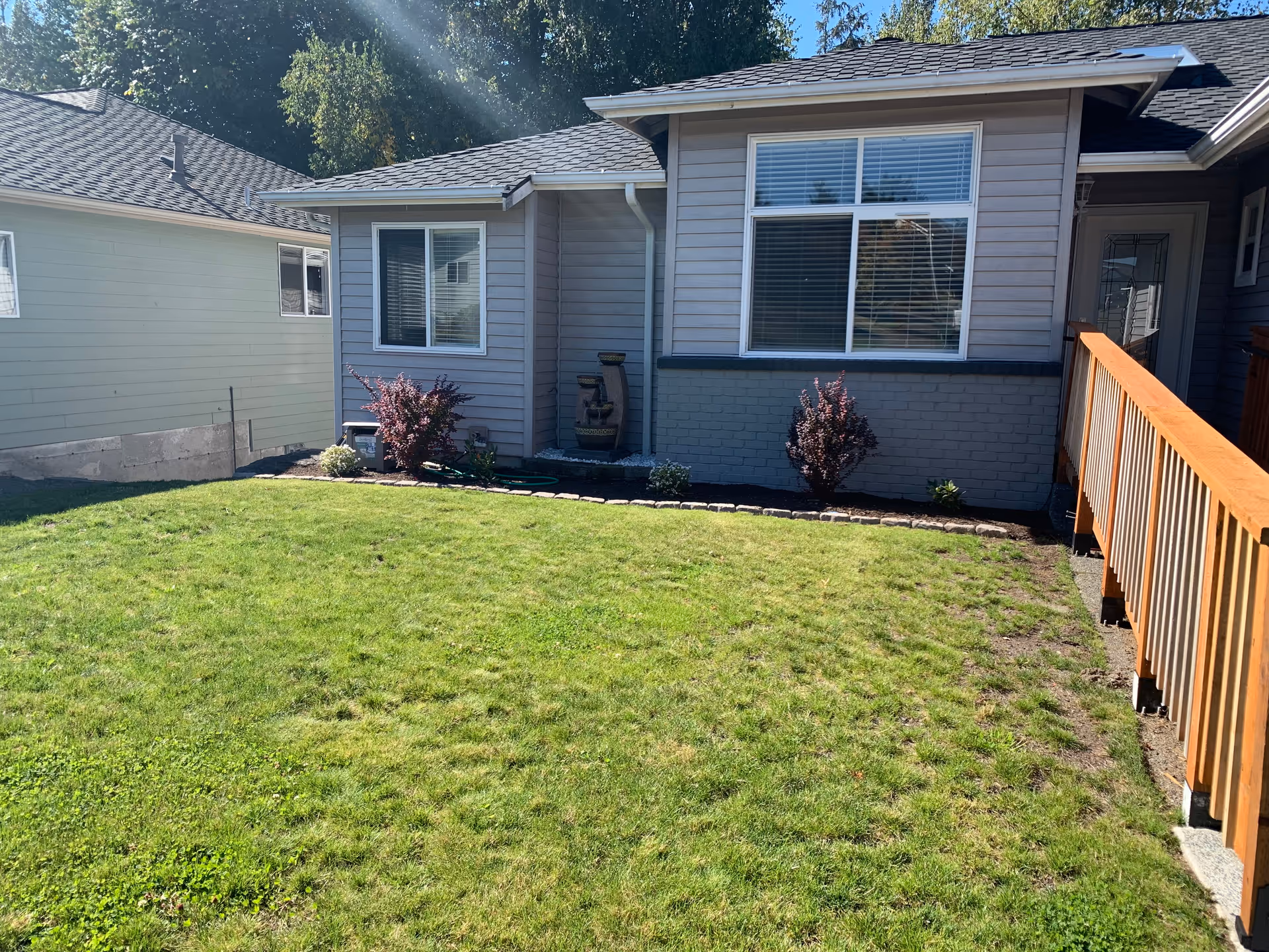 Front yard of a single-story house with a green lawn, small shrubs planted along the house's foundation, and a wooden handrail leading to the entrance door. The house has gray siding and large windows with white blinds.