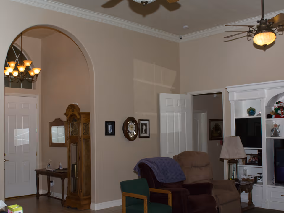 Interior view of a living room area with beige walls and a white ceiling. There is a brown recliner chair with a purple blanket draped over it, a green chair with wooden arms, a white entertainment center with shelves holding decorative items, and a lamp on a side table. A ceiling fan with a light fixture is visible. In the background, there is an arched doorway leading to a hallway with a wooden grandfather clock and a small table with items on it. A white door is also visible in the hallway.
