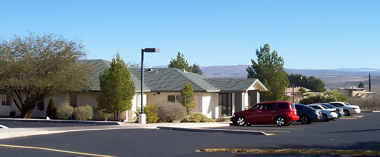 Front exterior of a single-story care facility with a parking lot, several cars, trees, and distant hills.