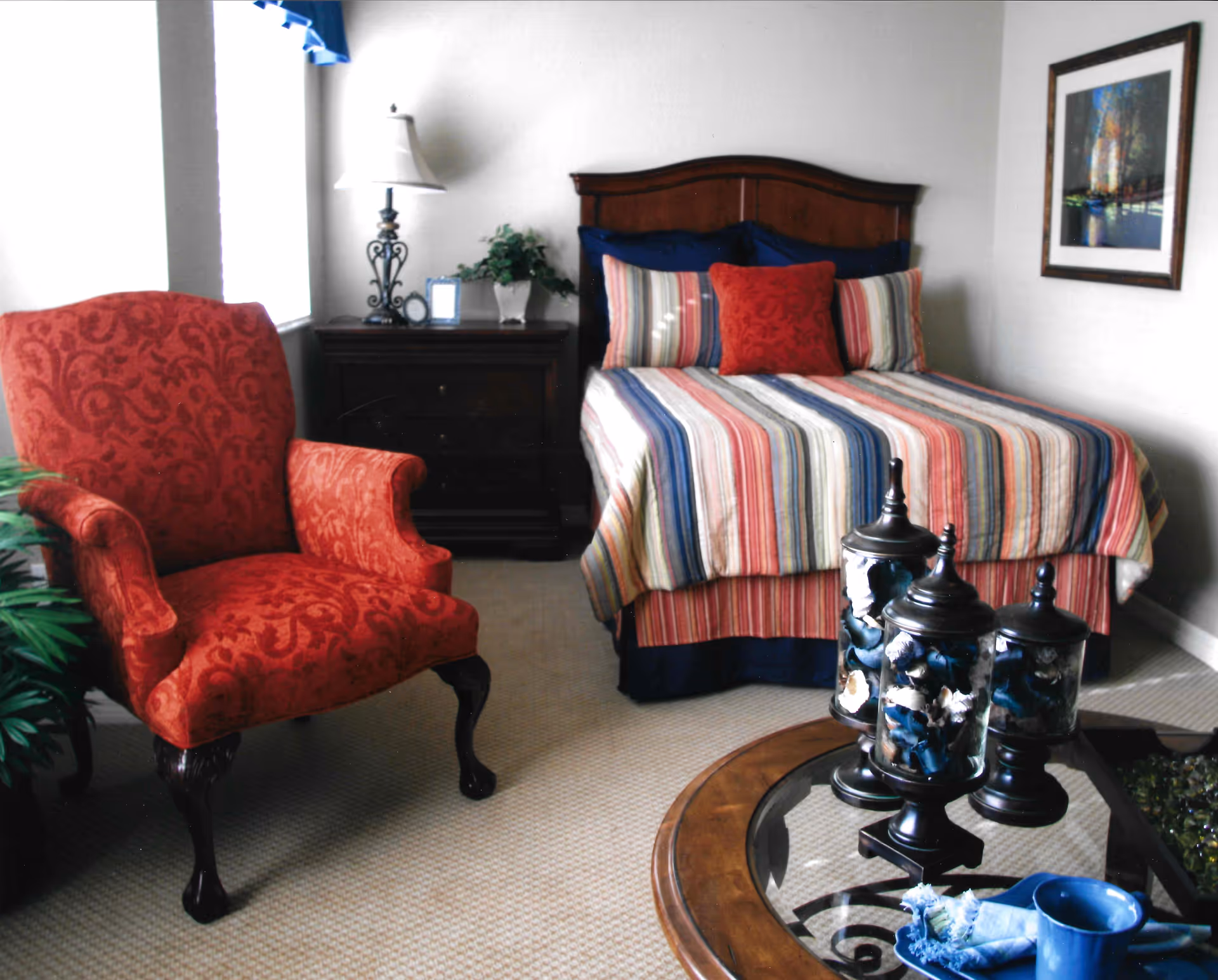 A cozy bedroom featuring a bed with a striped multicolored bedspread and matching pillows, a dark wooden nightstand with a lamp and a small plant, a red upholstered armchair with ornate wooden legs, and a round glass-top coffee table with decorative jars and a blue cup and napkin.
