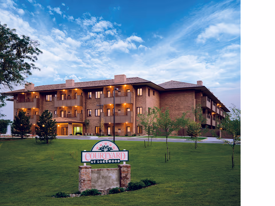 Exterior view of a three-story brick senior living facility building with balconies, surrounded by green grass and small trees under a partly cloudy blue sky. A sign in front reads 'Courtyard at Lakewood'.