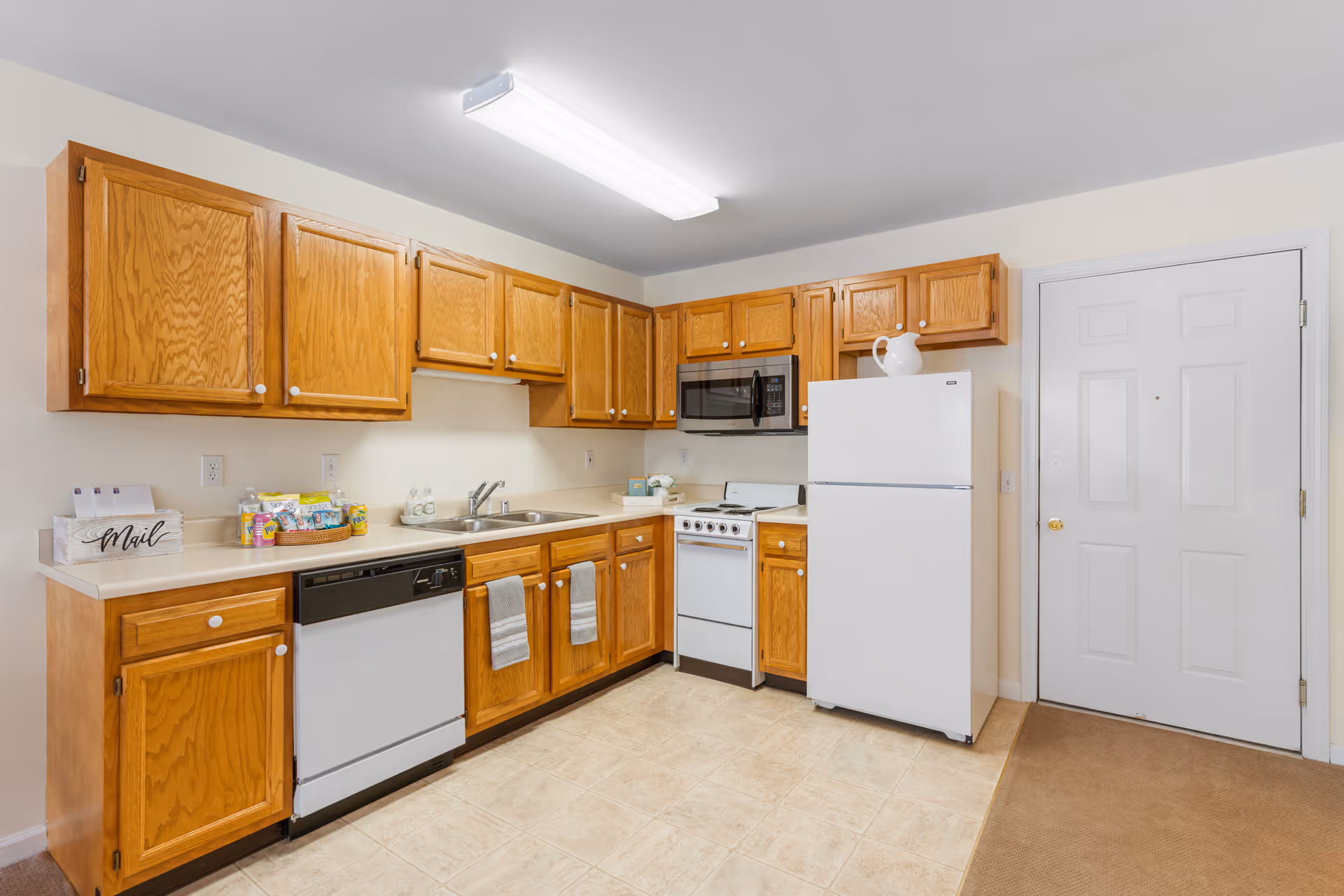 A clean and well-lit kitchen with wooden cabinets, a white refrigerator, a white stove with an overhead microwave, a dishwasher, and a double sink. The floor is tiled and there is a white door to the right. Various items such as a mail holder, canned drinks, and soap dispensers are placed on the countertops.
