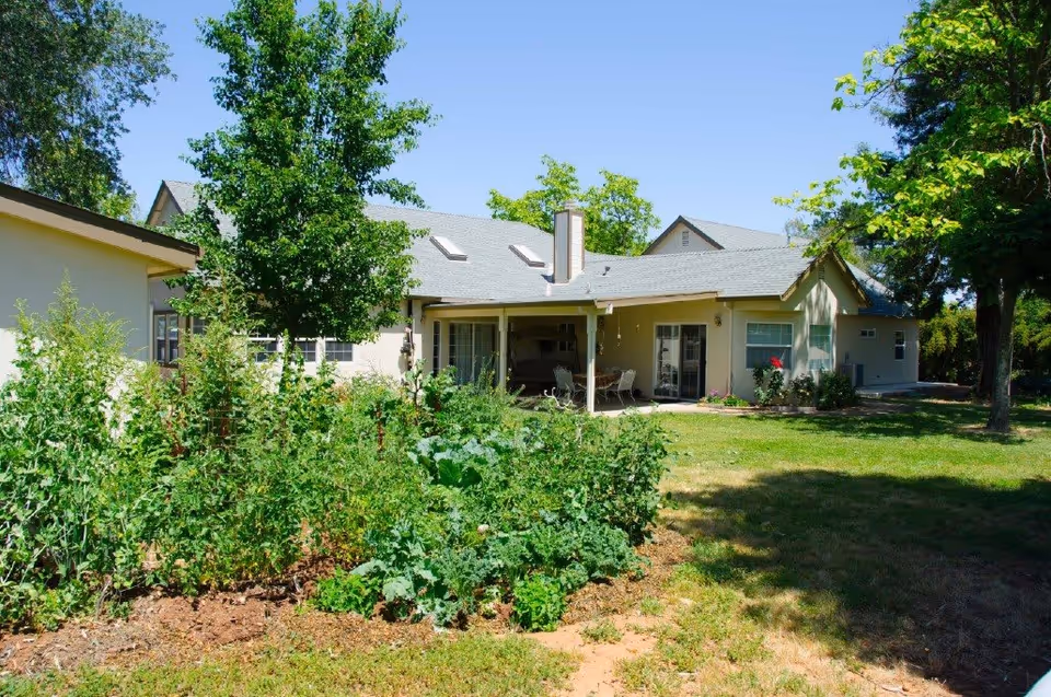 Single-story light-colored building with a covered patio, lawn and garden beds in the foreground under a clear blue sky.