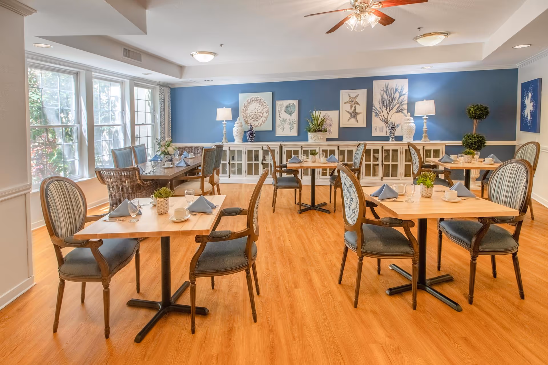 A bright dining room with wooden floors and multiple tables set with cups, glasses, and blue napkins. The room features elegant chairs with striped upholstery, large windows letting in natural light, blue accent walls decorated with framed sea-themed artwork, and lamps on a white sideboard along the back wall.