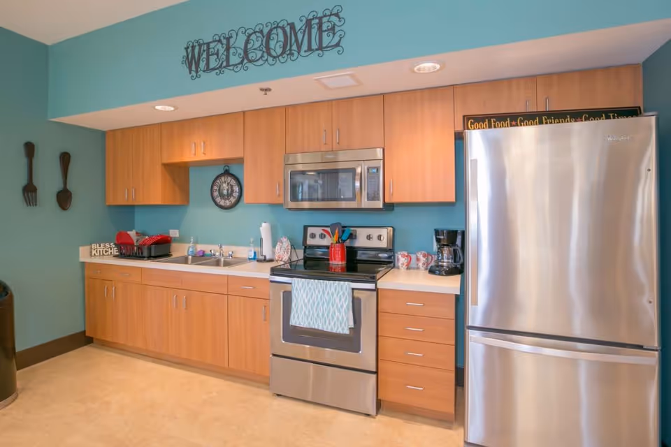 A modern kitchen with light wood cabinets, a stainless steel refrigerator, oven, and microwave. The walls are painted teal, and there is a decorative metal 'WELCOME' sign above the cabinets. On the counter, there is a coffee maker, mugs, a dish rack with red dishes, and various kitchen utensils. A clock is mounted on the wall above the sink.