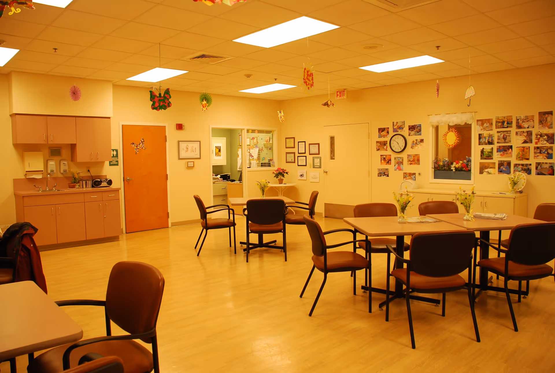 A brightly lit common room with several tables and brown chairs arranged around them. The walls are decorated with colorful hanging paper decorations and numerous photos. There is a small kitchenette area with cabinets, a sink, and a radio on the left side. A door with a restroom sign is visible, along with a clock on the wall and framed certificates. The floor is wooden, and the overall atmosphere is warm and welcoming.