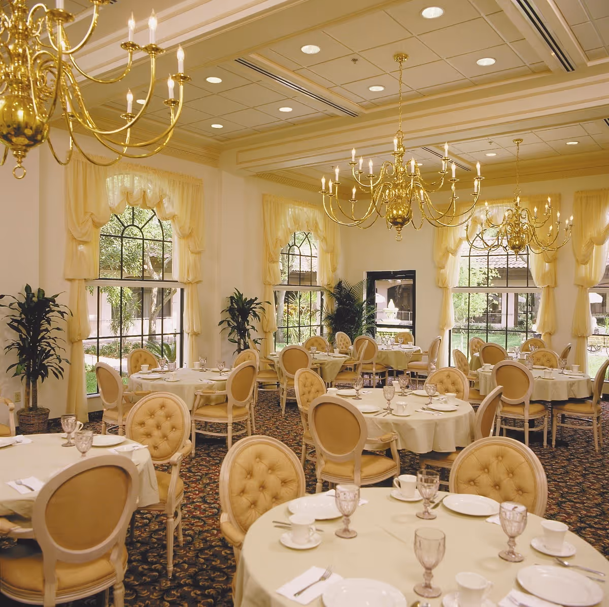 Elegant dining room with round tables covered in white tablecloths, set with plates, cups, and glasses. The room features large arched windows with cream-colored curtains, gold chandeliers hanging from the ceiling, and beige upholstered chairs. There are also green plants placed near the windows.