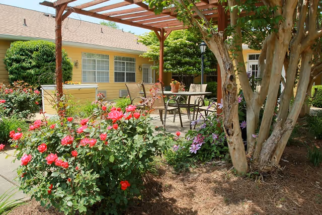 Outdoor patio area with a wooden pergola, metal table and chairs, surrounded by blooming red and purple flowers, trees, and shrubs in front of a yellow building with windows.