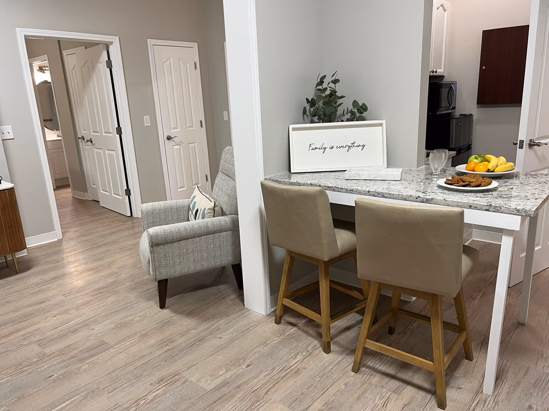Interior view of a senior living facility showing a small dining area with two beige chairs at a granite countertop table. On the table are a plate of cookies, a bowl of fruit, a glass of water, and a decorative sign that reads 'Family is everything.' To the left, there is a gray upholstered armchair with a patterned pillow. In the background, there are doors leading to other rooms, including a bathroom.