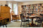 A cozy library room with wooden bookshelves filled with books along the wall, a wooden card catalog cabinet, a wicker chair by the window, and a round wooden table surrounded by black wooden chairs on a carpeted floor.