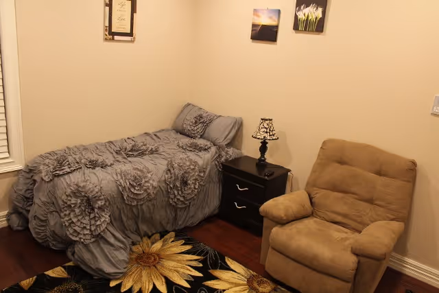 Small bedroom with a twin bed dressed in a gray ruffled comforter, a dark nightstand with a lamp, a beige recliner, and a floral rug.