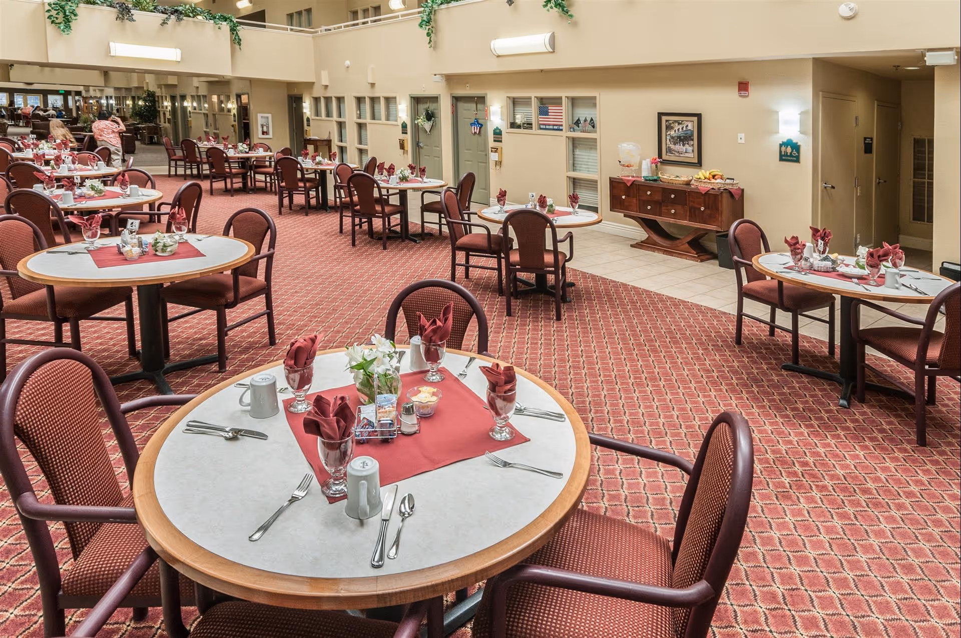 A spacious dining room in an assisted living facility with multiple round tables set with red napkins, glasses, silverware, and small flower arrangements. The room has a red patterned carpet, beige walls, and a sideboard with fruit and a beverage dispenser. There are a few people in the background near the entrance.