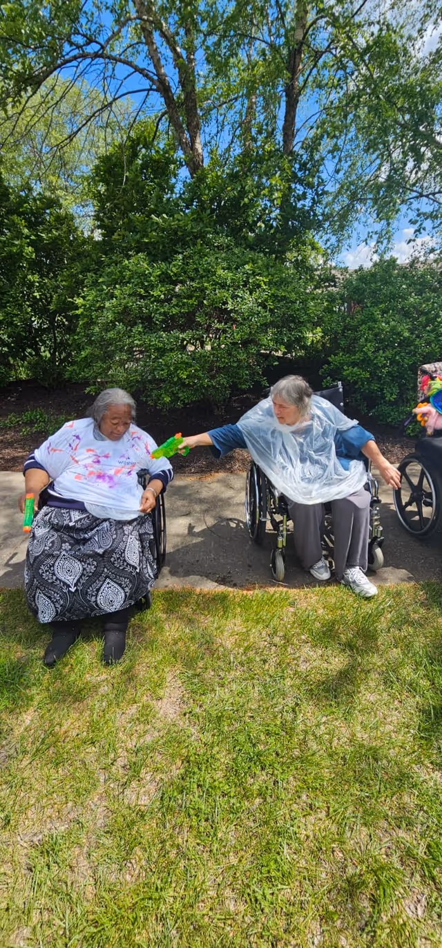Two elderly women in wheelchairs outdoors on a sunny day, playing with green and orange water guns. They are sitting on a paved path with green grass in front and lush green bushes and trees behind them. Both women are wearing plastic ponchos.