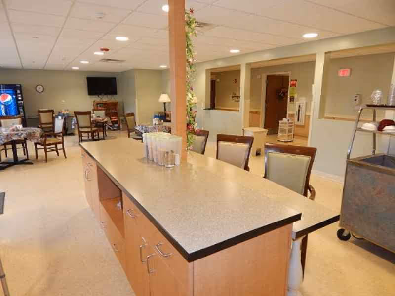 Interior view of a senior care facility dining area with a long counter in the foreground, several chairs around tables covered with tablecloths, a vending machine, a wall-mounted TV, and a serving cart on wheels.