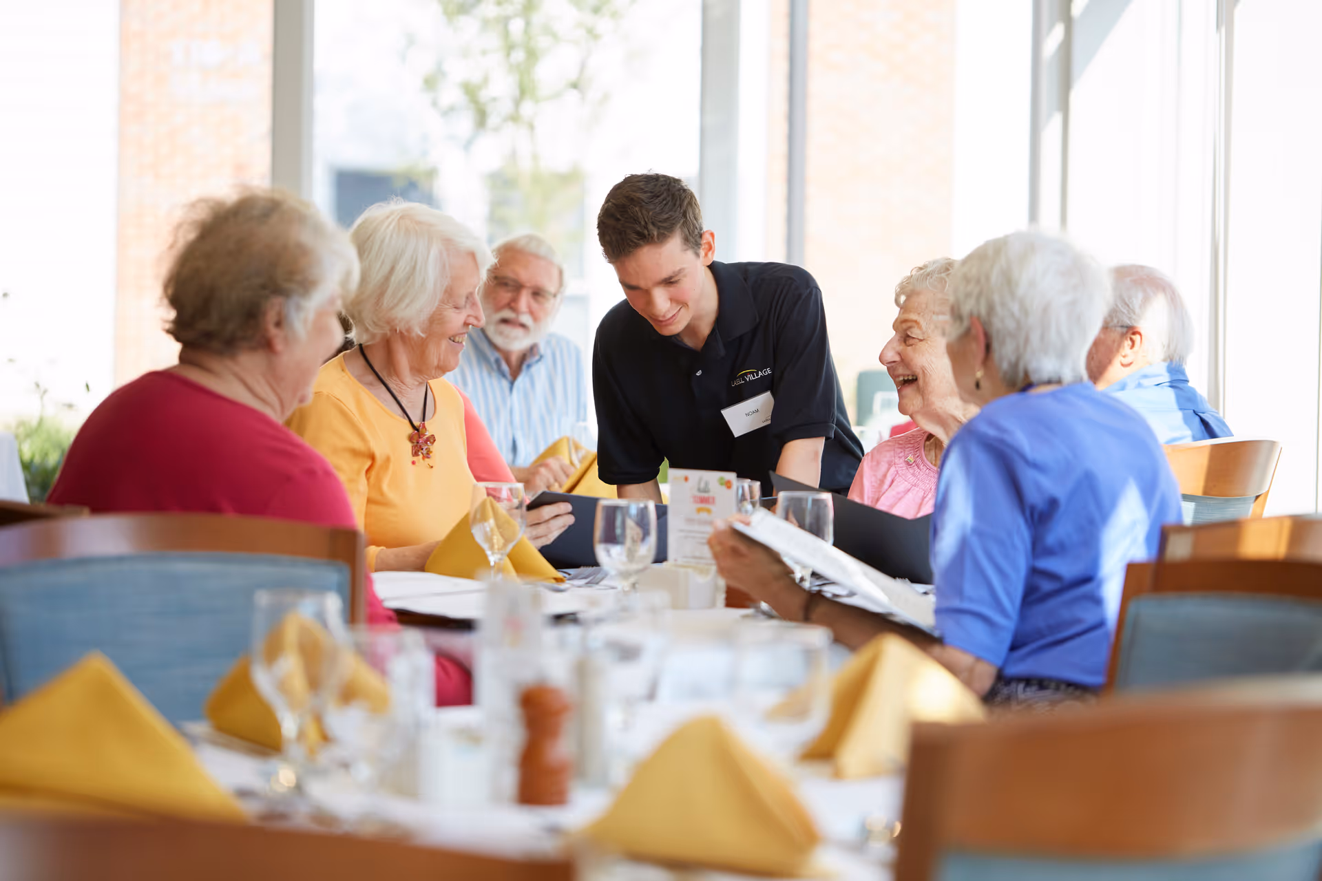 A group of elderly people sitting around a dining table with yellow napkins and glassware, interacting with a young male staff member wearing a black shirt with a Lasell Village name tag, in a bright room with large windows.