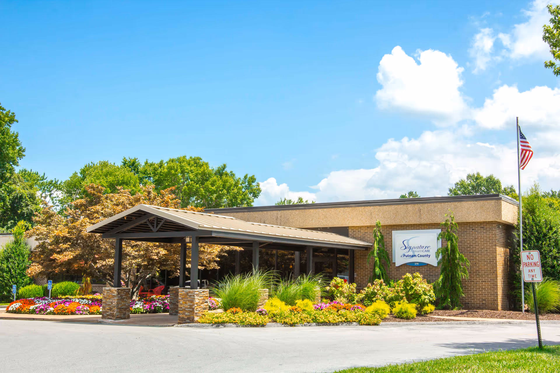 Exterior view of Signature HealthCARE of Putnam County building with a covered entrance, landscaped flower beds, green trees, and an American flag on a flagpole under a blue sky with some clouds.
