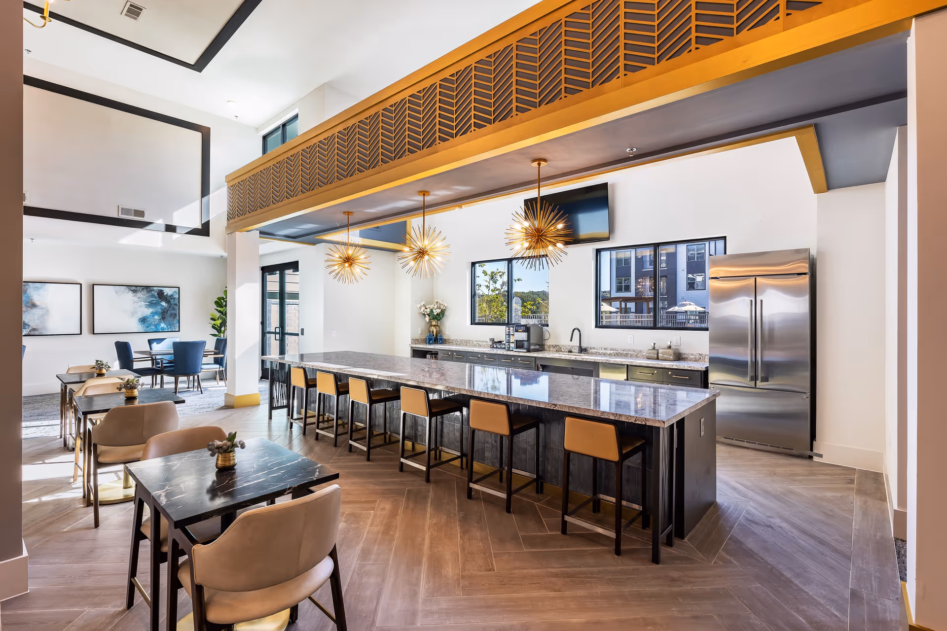 A modern communal kitchen and dining area with a long marble island countertop surrounded by bar stools. The kitchen features stainless steel appliances, including a refrigerator, and a coffee machine on the counter. The space is well-lit with large windows and decorative pendant lights hanging above the island. There are several small dining tables with chairs and decorative plants, and the room has a high ceiling with a decorative wooden beam.