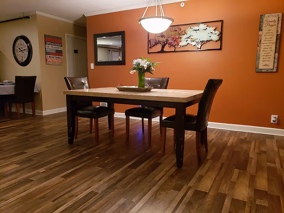 A dining room with a rectangular wooden table and four dark brown leather chairs. A green vase with flowers is placed on the table. The walls are painted orange and beige, decorated with a large metal tree wall art, a rectangular mirror, a clock, and framed inspirational quotes. The floor is wooden with a polished finish, and a hanging light fixture illuminates the table.