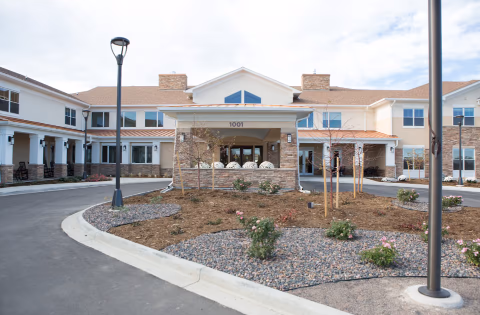 Front exterior view of a two-story assisted living facility with a covered entrance, stone and beige siding, multiple windows, landscaped garden with small plants and flowers, and street lamps along the driveway.