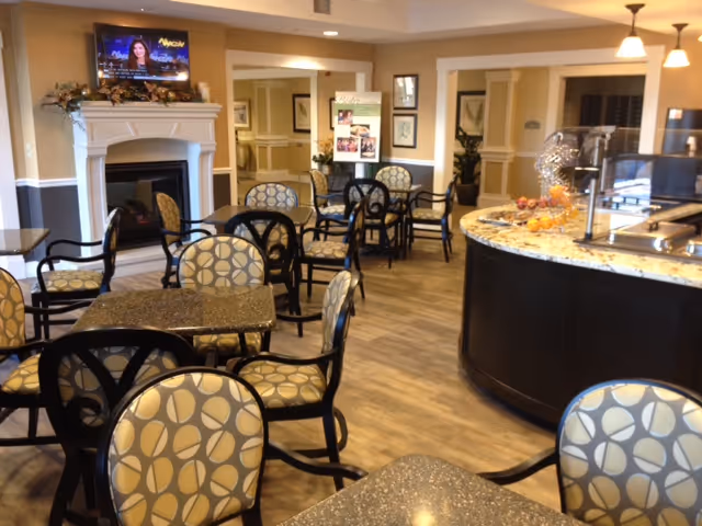 An elegant senior dining room with patterned chairs and tables, a curved serving counter, fireplace and a wall-mounted TV.