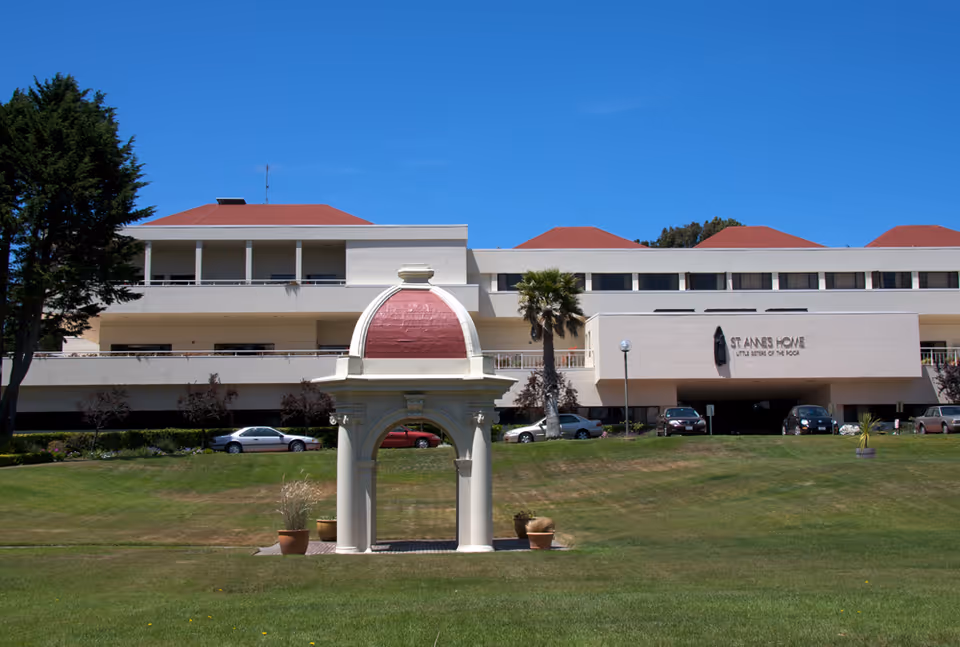 Exterior view of St. Anne's Home For The Aged building with a manicured lawn and a small decorative pavilion with a red dome in the foreground under a clear blue sky.