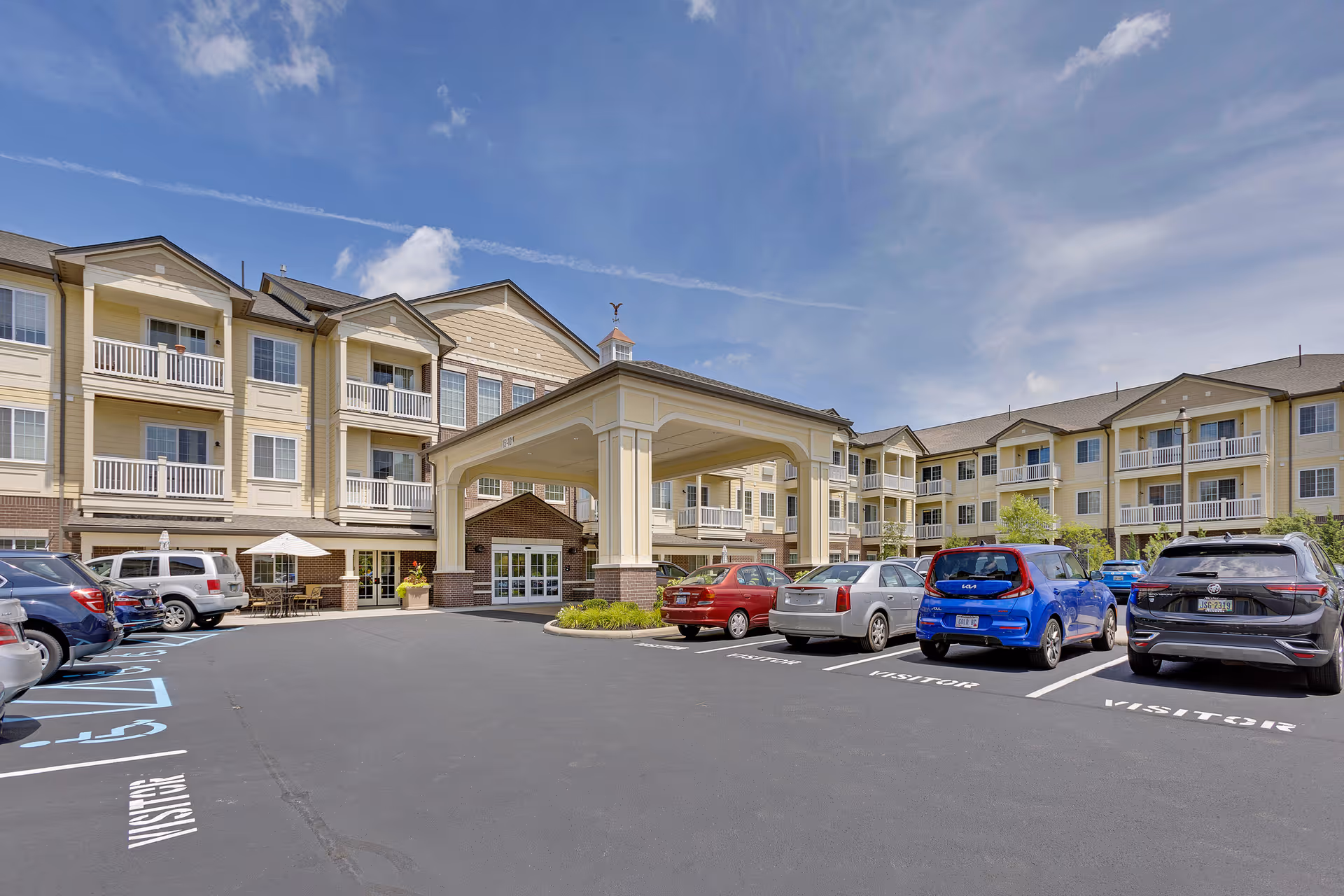 Exterior view of a multi-story senior living facility with beige and brick facade, balconies, and a covered entrance. Several cars are parked in the visitor parking spaces in front of the building under a partly cloudy sky.