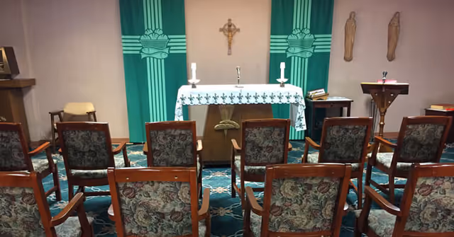 Small chapel with an altar, crucifix, candles, lectern and rows of upholstered chairs facing forward.