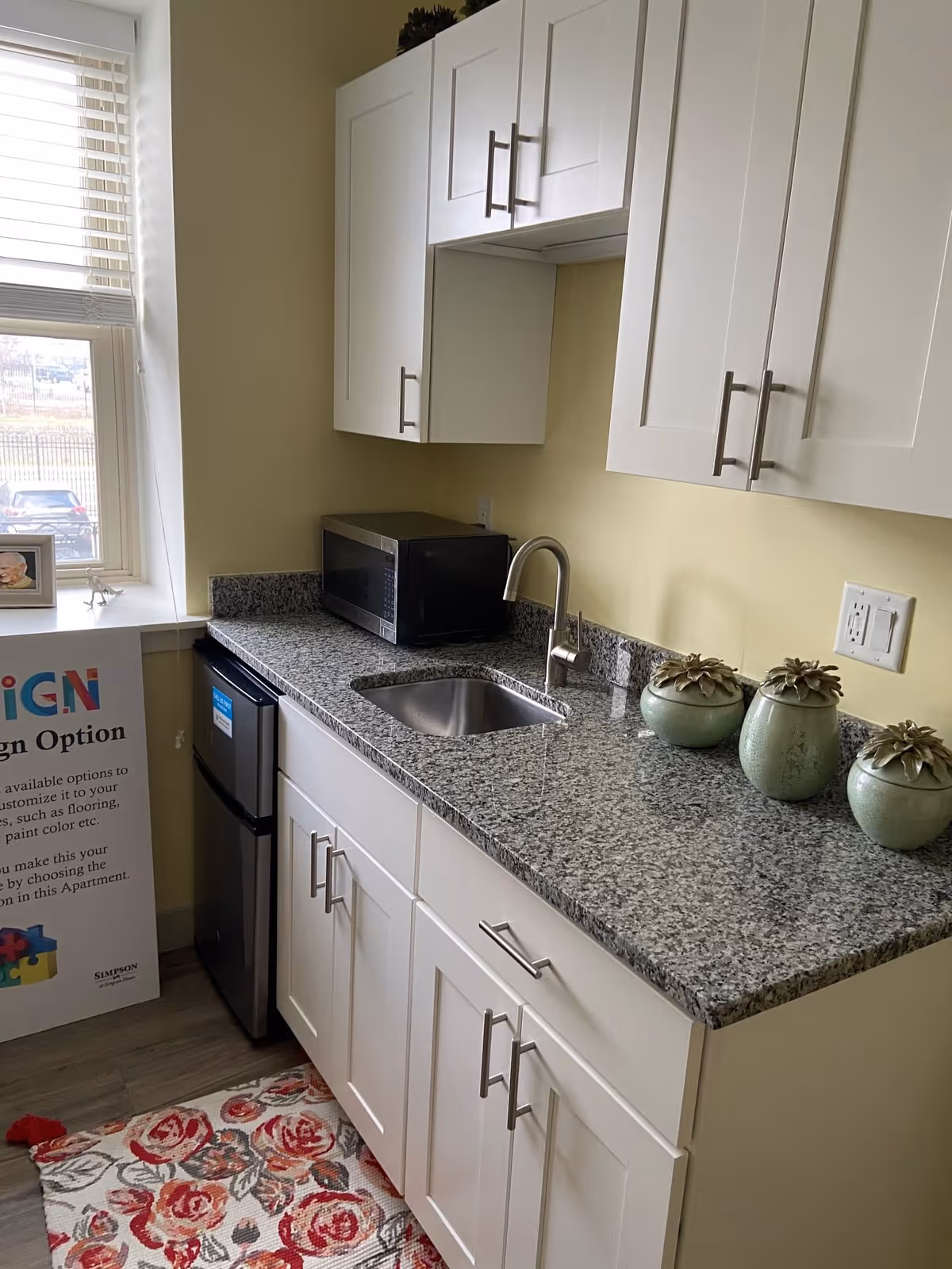 A small kitchen area with white cabinets, a granite countertop, a stainless steel sink with a modern faucet, a microwave, and a mini refrigerator. There are three green decorative pots on the counter and a floral patterned rug on the floor. A window with blinds is on the left side, and a sign with colorful text is partially visible near the window.