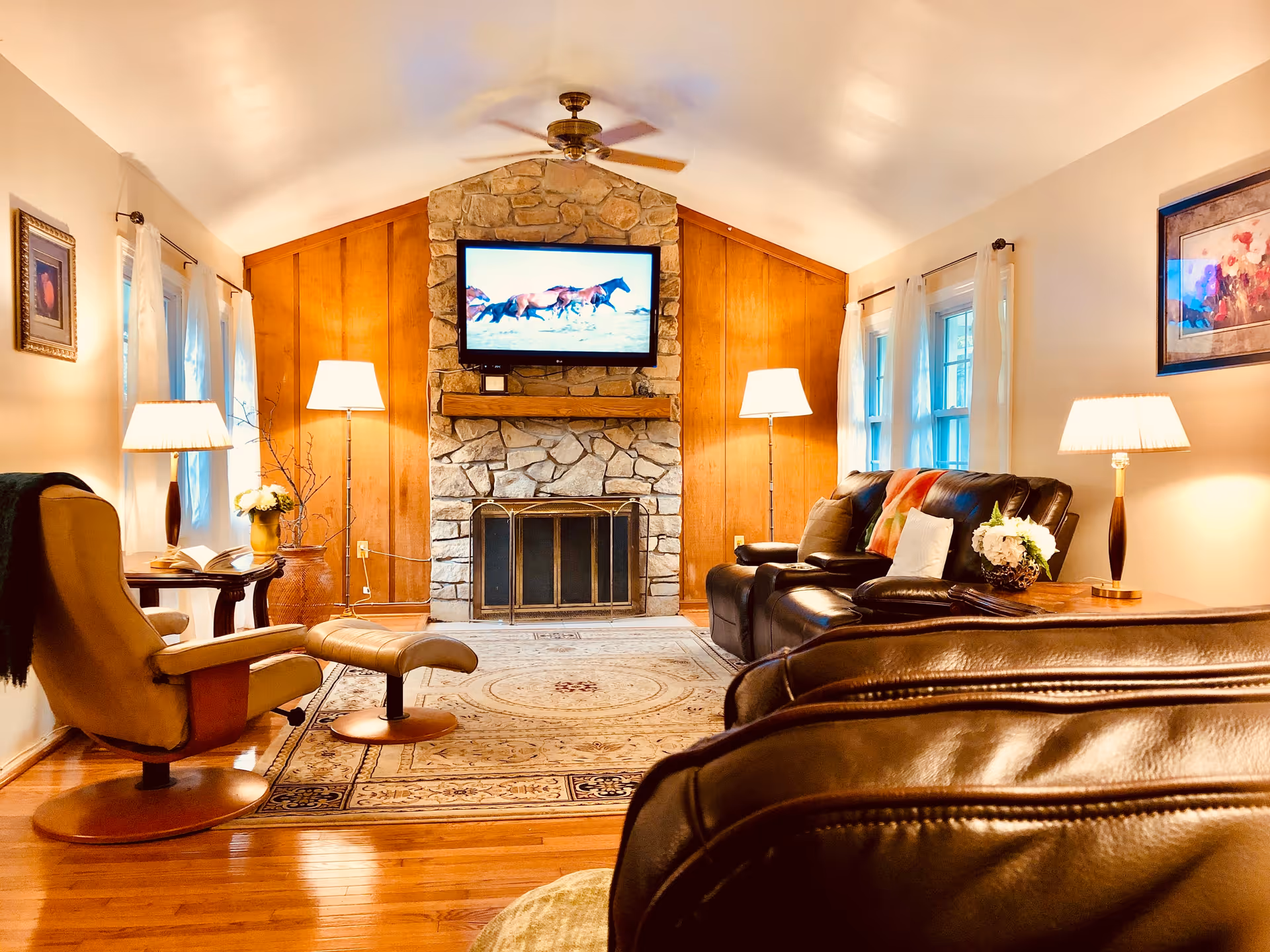 A cozy living room with a stone fireplace and a mounted flat-screen TV displaying running horses. The room features wooden paneling on the far wall, hardwood floors, and a large area rug. There are two leather recliners, a cushioned armchair with a matching ottoman, and several floor and table lamps providing warm lighting. Windows with sheer curtains line the walls, and framed artwork is hung on the walls.