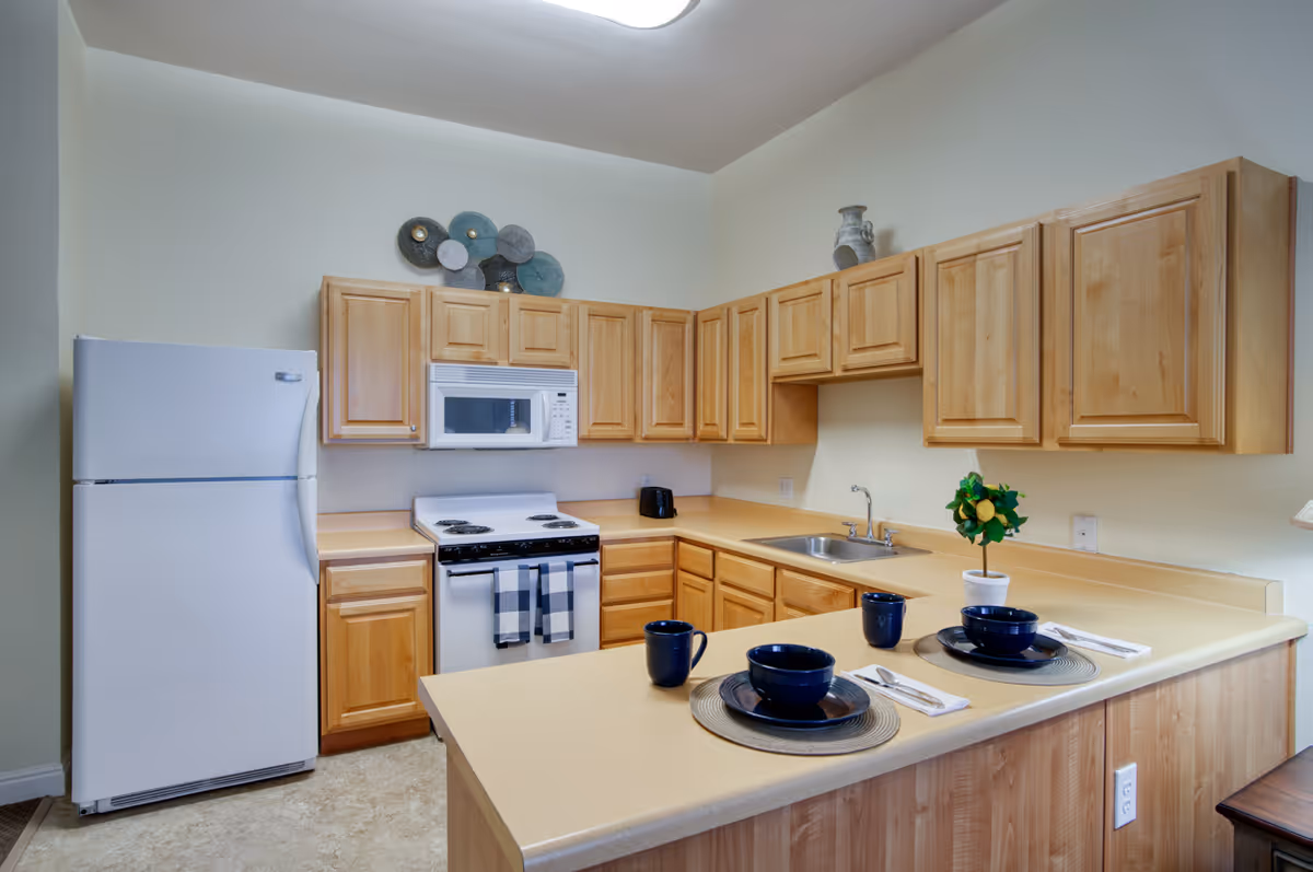 A bright kitchen with light wood cabinets, a white refrigerator, stove with oven, microwave, and a sink. The countertop has two place settings with blue bowls and mugs, silverware on napkins, and a small potted plant with green leaves and yellow fruit. Decorative wall art is above the cabinets.