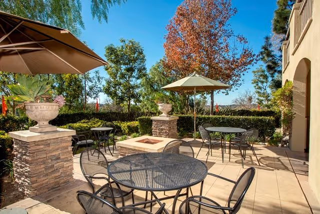 Sunlit outdoor patio with metal tables and chairs, umbrellas, planters, and a central square fire pit surrounded by trees and hedges.