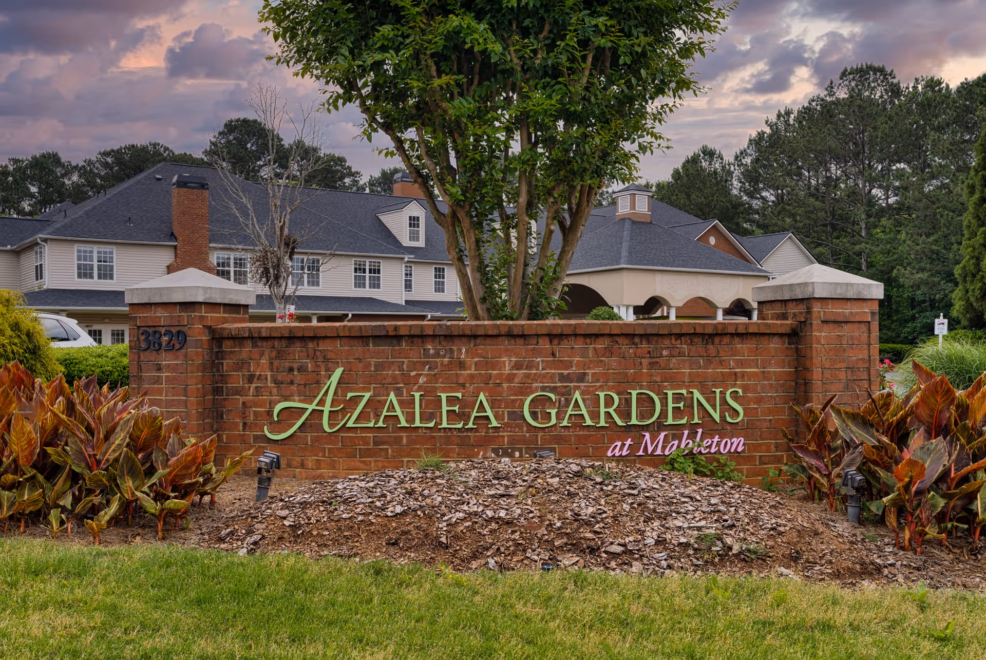 Brick entrance sign for Azalea Gardens at Mableton with green and purple lettering, surrounded by plants and trees, with a large building and cloudy sky in the background.
