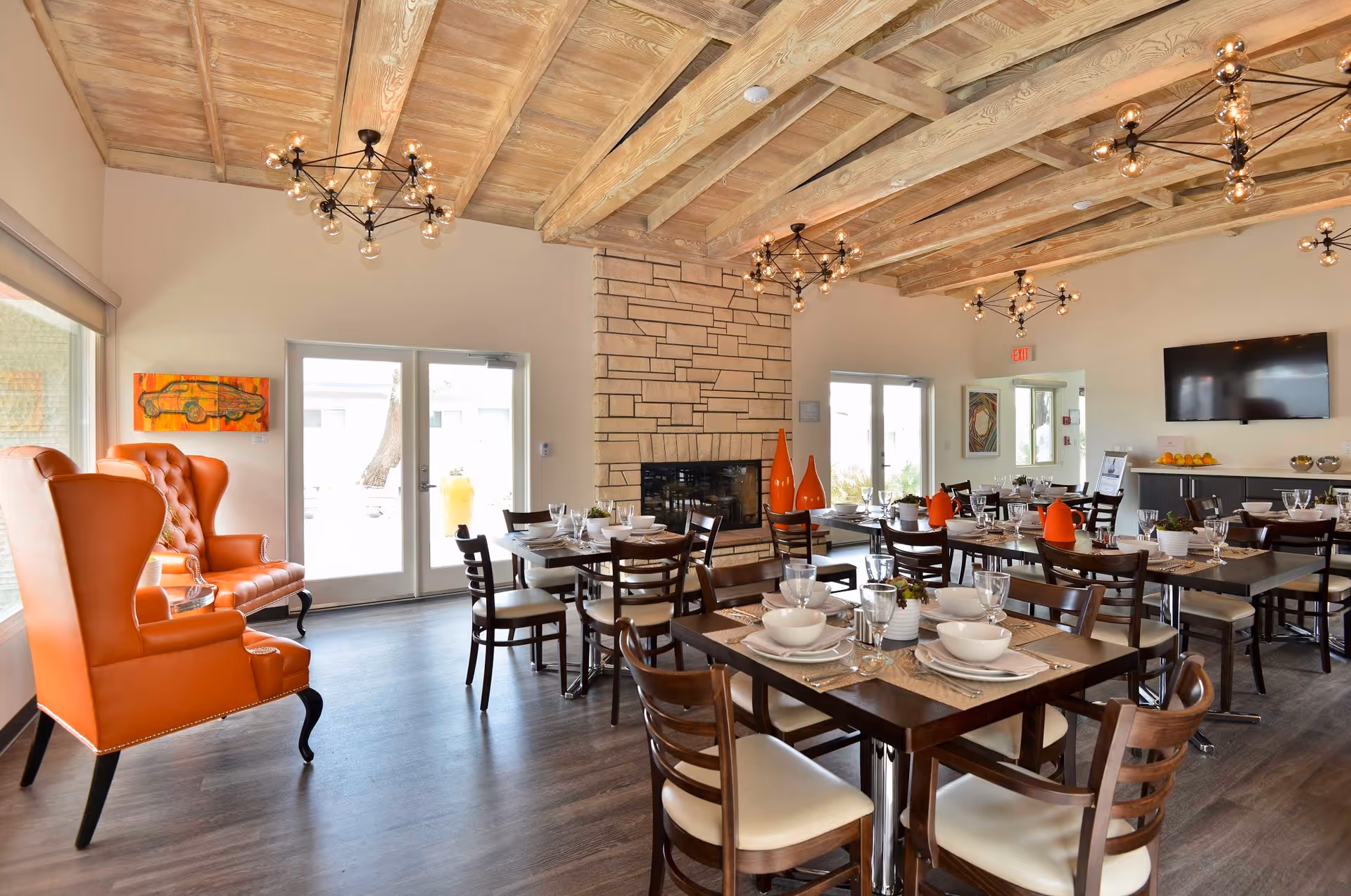 A bright dining room with multiple wooden tables set with white bowls, plates, and glasses. The room features a stone fireplace in the center, wooden ceiling beams, and modern light fixtures. There are orange high-back chairs near the window and orange decorative vases on some tables. A flat-screen TV is mounted on the wall above a sideboard with fruit and other items.