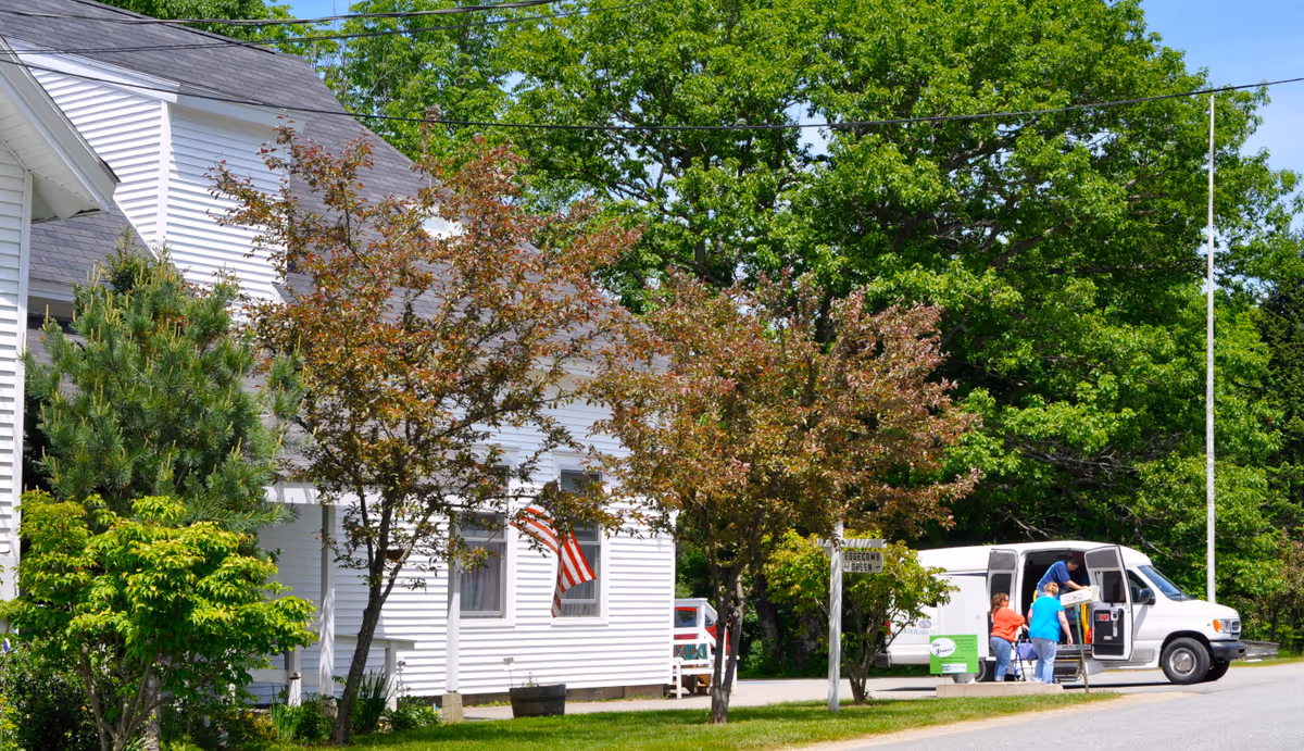 White siding building with an American flag, trees and shrubs in the yard, and a white van with people parked at the curb.