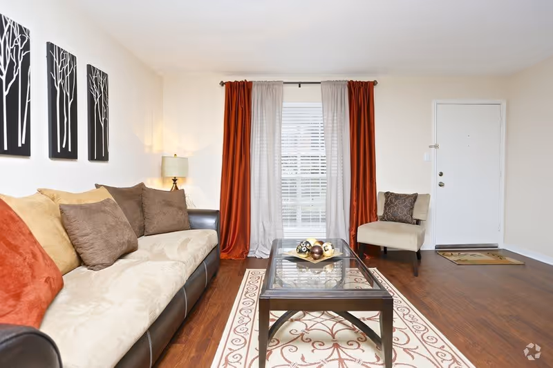 A bright living room with a beige and brown sofa adorned with multiple cushions, a glass coffee table on a patterned rug, a beige armchair with a decorative pillow, a floor lamp, and a window with red and white curtains. The room has wooden flooring and a white door near the armchair.
