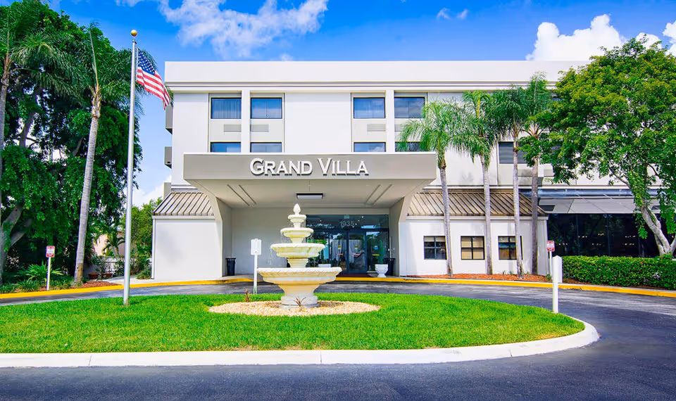 Front exterior view of the Grand Villa building with a three-tiered fountain in the circular driveway, an American flag on a flagpole to the left, palm trees and other greenery surrounding the entrance, and a clear blue sky above.