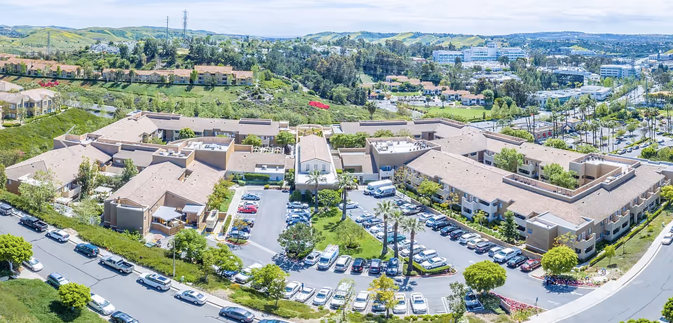 Aerial view of Heritage Pointe senior living facility showing multiple beige buildings with brown roofs arranged around parking lots filled with cars. The facility is surrounded by green hills and trees, with a cityscape visible in the background under a partly cloudy sky.