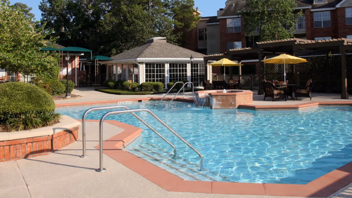 Outdoor swimming pool area with clear blue water, metal handrails for pool entry, a hot tub with water flowing into the pool, patio tables with yellow umbrellas, and a building with large windows and greenery surrounding the area.