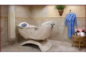 A modern white hydrotherapy bathing tub in a tiled bathroom with a blue robe hanging and a small wicker table holding towels and flowers.