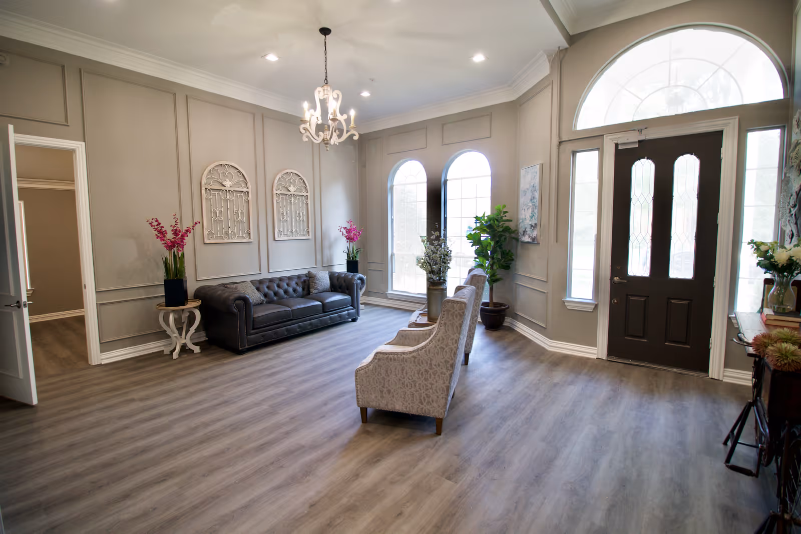 A bright and spacious living room area with light gray walls and wood flooring. The room features a dark leather tufted sofa, two patterned armchairs, a chandelier hanging from the ceiling, and large arched windows letting in natural light. There are decorative wall hangings, potted plants, and a dark front door with glass panels.