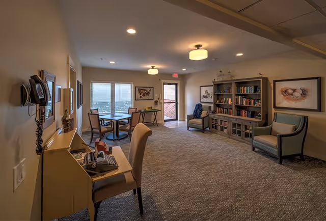 A cozy senior living common area with carpeted floor, beige walls, and ceiling lights. The room features a small desk with a chair and telephone, a round table with four chairs near a window, two armchairs, and a large bookshelf filled with books. Framed artwork is displayed on the walls, and a door leads outside.