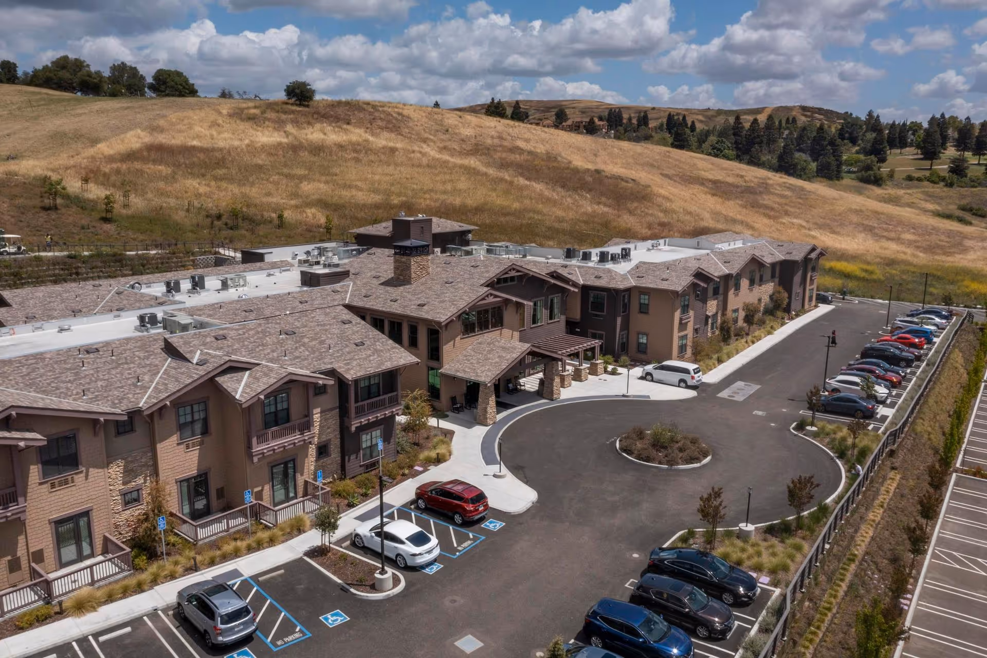 Aerial view of Discovery Commons San Ramon, showing a large two-story senior living facility with a circular driveway and multiple parked cars. The building is surrounded by landscaped areas and dry grassy hills in the background under a partly cloudy sky.