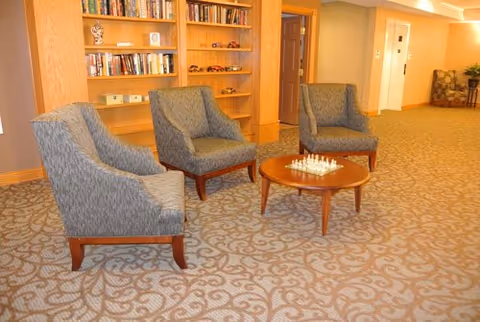 A cozy seating area in a senior living facility featuring three upholstered armchairs arranged around a small wooden round table with a chess set on it. Behind the chairs is a built-in wooden bookshelf filled with books and decorative items. The room has patterned carpet flooring and warm lighting.