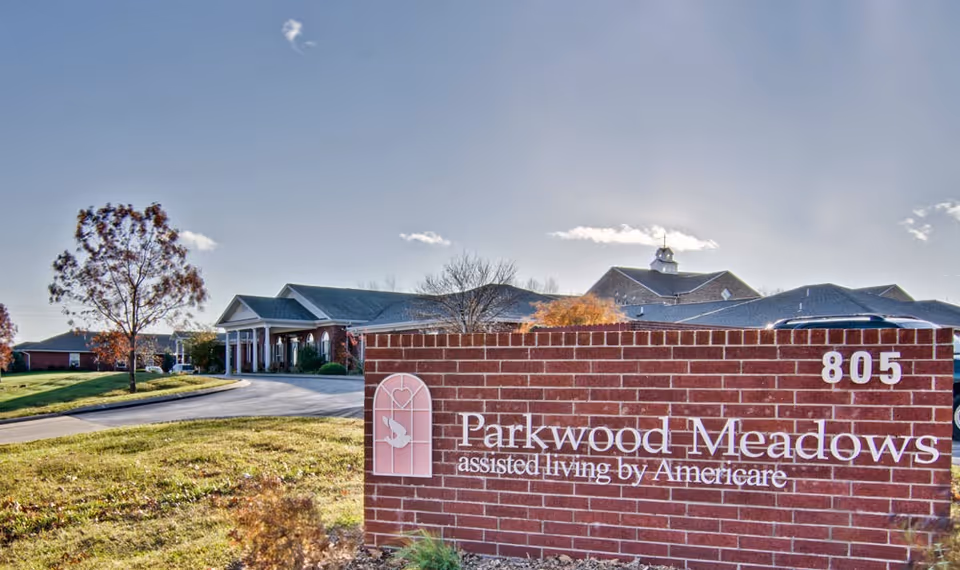 Exterior view of Parkwood Meadows Senior Living facility with a brick sign in the foreground displaying the name and address number 805. The building and driveway are visible in the background under a clear sky.