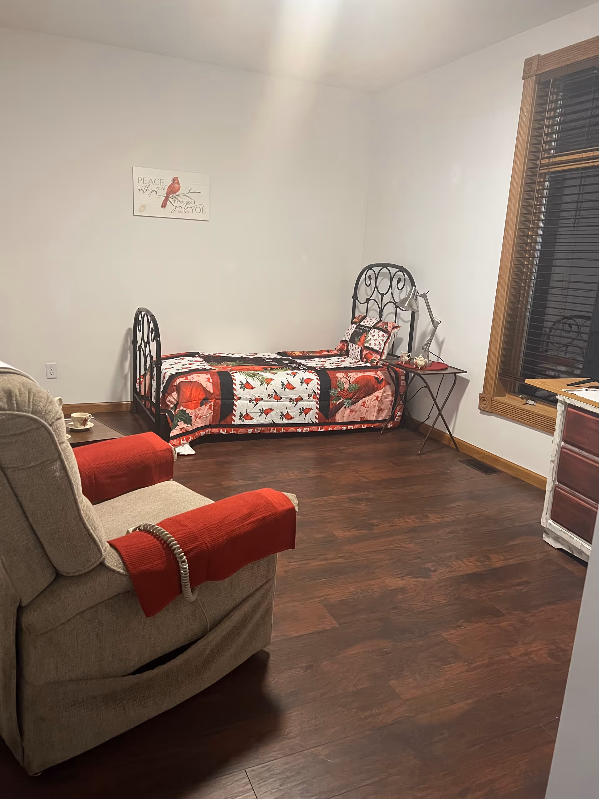 A cozy bedroom with a single bed featuring a red and white patterned quilt and matching pillow. Next to the bed is a small side table with a lamp. A beige recliner chair with red armrest covers is positioned near the bed. The room has wooden flooring, white walls, and a window with wooden blinds. A small framed artwork with a bird and text hangs on the wall above the bed.