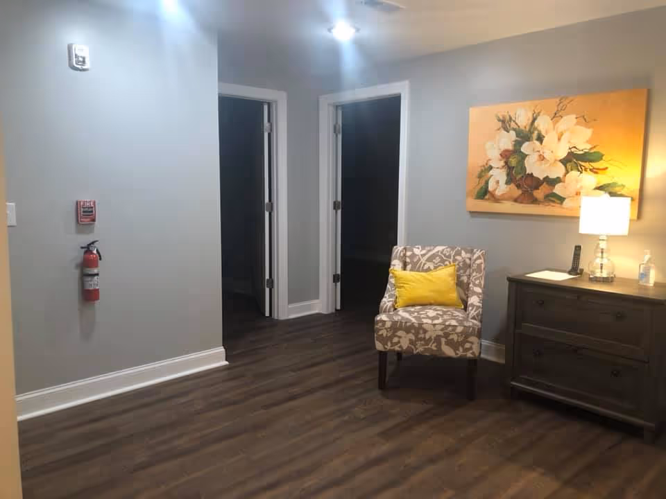 Small seating area in a senior living facility hallway with a patterned chair, yellow pillow, side table and lamp, floral painting, and two open doorways.