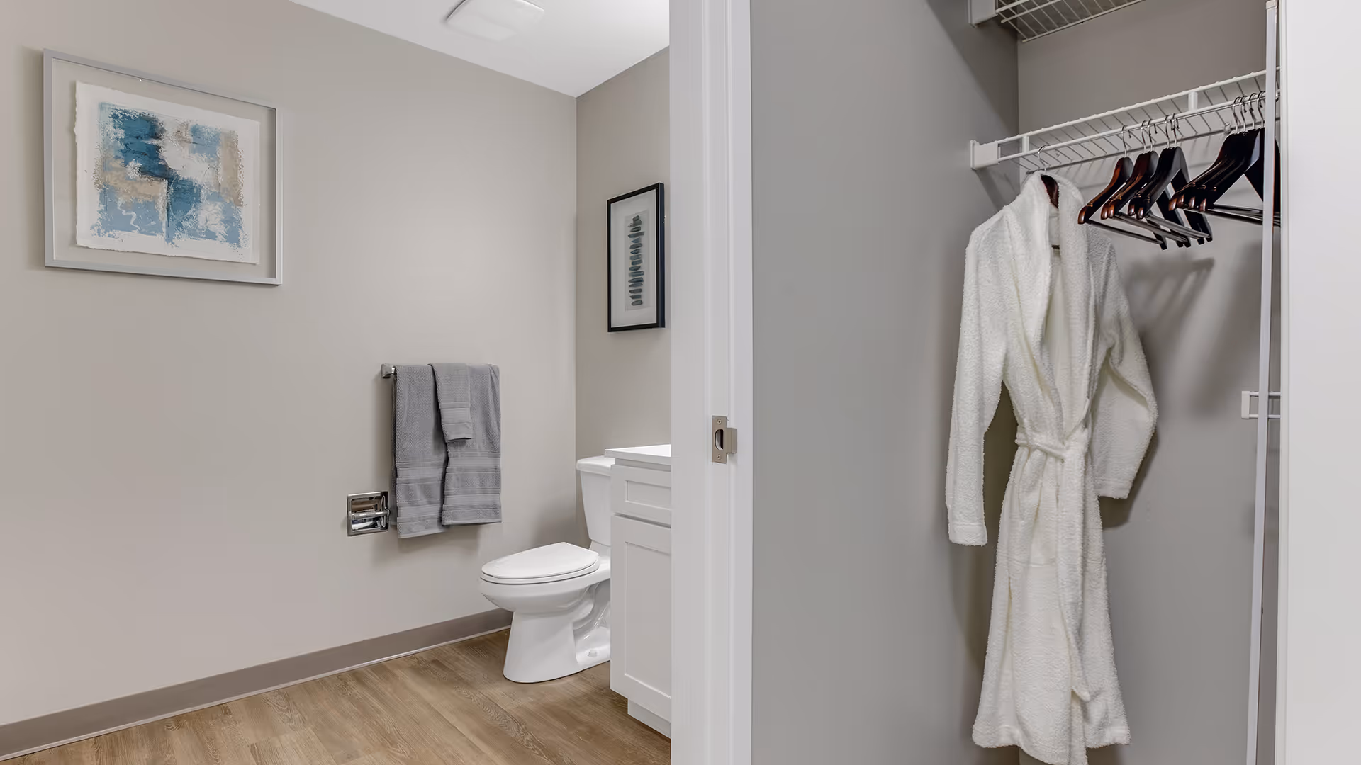 A clean bathroom with a white toilet, a white vanity with a sink, and two gray towels hanging on a wall-mounted towel rack. There is a framed abstract painting on the wall above the towels. To the right, an open closet with a white bathrobe hanging on a hanger and several empty hangers on a wire shelf.