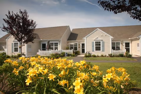 Single-story beige building front with a bed of yellow lilies, small white picket fences, and a paved walkway.